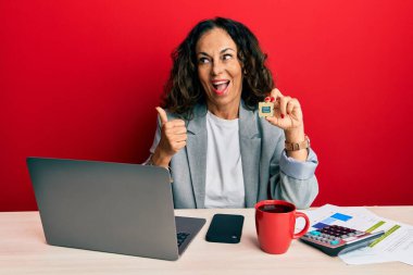 Güzel orta yaşlı bir kadın bilgisayar işlemcisini elinde tutuyor başparmağıyla yan tarafa bakıyor ve ağzı açık gülümsüyor. 