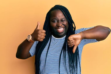 Örgülü genç siyahi kadın baş parmağını kaldırıyor ve kameraya seksi bir ifadeyle bakıyor, neşeli ve mutlu bir yüzle.. 