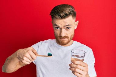 Genç kızıl saçlı adam diş fırçasını diş macunuyla tutuyor ve bir bardak su şüphecisi ve gergin, sorun yüzünden suratını asıyor. negatif kişi. 