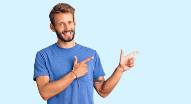 Yakışıklı, sakallı, sıradan kıyafetler giyen, gülümseyen ve kameraya bakan iki eli ve parmakları yanda olan sarışın bir adam.. 