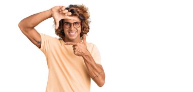 Gündelik kıyafetler ve gözlük takan genç İspanyol adam gülümsüyor ve mutlu yüzlü eller ve parmaklarla çerçeve yapıyor. yaratıcılık ve fotoğrafçılık kavramı. 