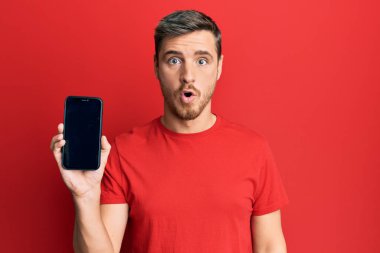 Akıllı telefon tutan yakışıklı beyaz adam ekranda korkmuş ve ağzı açık bir şekilde şaşkınlık ve şaşkınlık dolu bir ifade sergiliyor. 