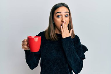 Genç esmer kız bir fincan kahve içiyor ağzını eliyle kapatıyor, şokta ve hata yapmaktan korkuyor. şaşırmış ifade 