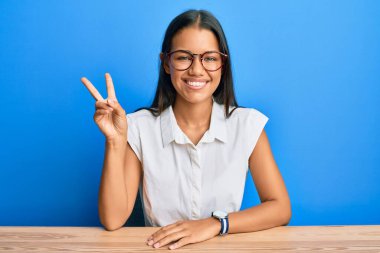 Güzel İspanyol kadın, gündelik giysiler içinde masada oturuyor ve iki numaralı parmaklarını göstererek gülümsüyor. Kendinden emin ve mutlu.. 