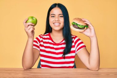 Genç Asyalı kız hamburgerle sağlıklı elmayı karşılaştırıyor mutlu ve havalı bir gülümsemeyle gülümsüyor. diş gösterme. 