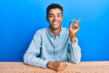 Genç, yakışıklı, Afro-Amerikalı, gündelik giysiler giymiş, yüzünde kocaman bir gülümsemeyle masada oturuyor, kameraya el ile işaret ediyor.. 