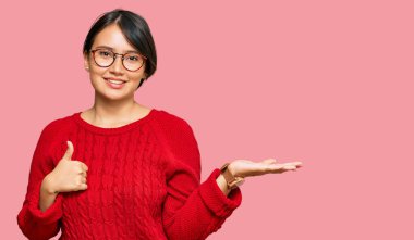 Kısa saçlı, günlük süveter giyen, avuçlarını gösteren ve baş parmağıyla hareket eden mutlu ve neşeli gülümseyen genç, İspanyol bir kadın. 