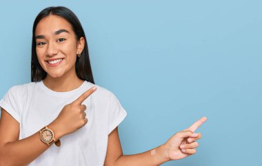 Genç Asyalı kadın, günlük beyaz tişört giyiyor gülümsüyor ve kameraya bakıyor. İki eliyle ve parmaklarıyla kenarı işaret ediyor.. 