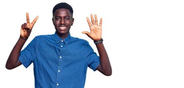 Genç, Afro-Amerikalı, günlük giysiler giymiş, 8 numaralı parmaklarıyla işaret ederken, kendinden emin ve mutlu bir şekilde gülümsüyor.. 