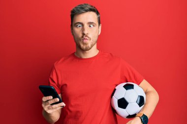 Futbol topu tutan yakışıklı beyaz adam akıllı telefona bakıyor ağzıyla balık suratı yapıyor ve şaşı bakıyor, çılgın ve komik.. 