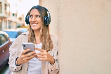 Beyaz kadın şehirde akıllı telefon ve kulaklık kullanarak mutlu bir şekilde gülümsüyor..