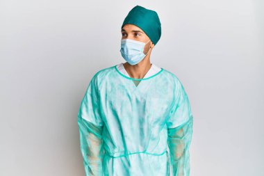 Cerrah üniforması ve tıbbi maske takan genç yakışıklı adam gülümseyip yan tarafa bakıyor ve düşünüyor.. 