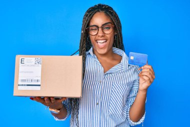 Saçları örgülü ve kredi kartı takılı genç Afro-Amerikalı kadın kameraya seksi bir ifadeyle bakıyor, neşeli ve mutlu bir yüzle.. 