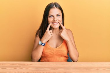 Gündelik giysiler giymiş genç beyaz kadın masaya oturmuş ağzı açık gülümsüyor, parmakları işaret ediyor ve zorla neşeli bir gülümseme sergiliyor. 