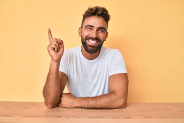 Latin asıllı genç adam gündelik giysiler içinde masaya oturmuş gülümsüyor mutlu bir şekilde elini ve parmağını yana doğru uzatıyordu. 