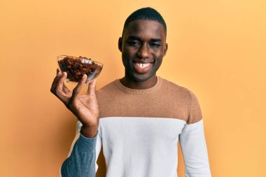 Afro-Amerikalı genç adam elinde kuru üzümlü bir kaseyle pozitif ve mutlu görünüyor ve kendine güvenen bir gülümsemeyle dişlerini gösteriyor. 