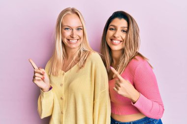Pembe arka planda yan yana duran iki arkadaş gülümsüyor ve kameraya bakıyor iki eliyle ve parmaklarıyla kenarı işaret ediyor.. 