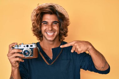 Şapkalı genç İspanyol adam elinde klasik kamera, gülümsüyor mutlu bir şekilde eli ve parmağı işaret ediyor. 