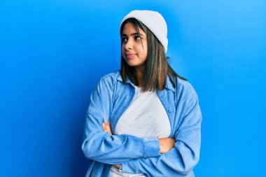 Şirin yün şapkalı genç Latin kadın kollarını kavuşturup yan tarafa bakıyor ikna olmuş ve kendinden emin. 