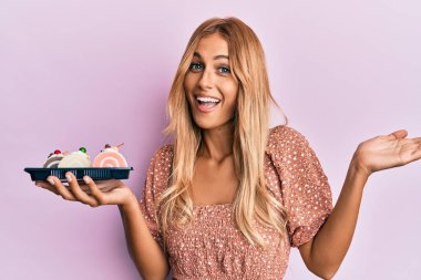 Güzel sarışın genç kadın pasta tatlısı tutarak başarıyı mutlu bir gülümsemeyle ve kazanan yüz ifadesiyle kutluyor. 