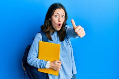 Genç esmer kız öğrenci çantası takıyor. Başparmak kaldırıyor. Korkmuş ve şaşırmış bir şekilde jest yapıyor.. 