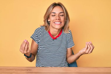 Güzel, beyaz bir kadın, gündelik giysiler içinde masada mutlu ve heyecanlı bir şekilde oturuyor. Kolları havada, gülümsüyor ve başarı için bağırıyor. kutlama konsepti. 