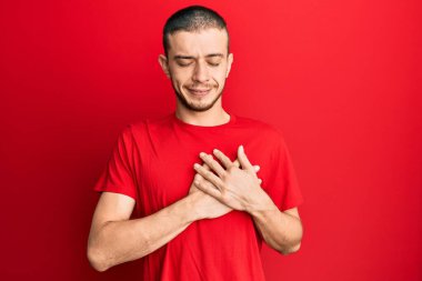 Normal kırmızı tişört giyen İspanyol genç adam elleri göğsünde gülümsüyor, gözleri kapalı ve yüzünde minnet dolu bir jest var. sağlık kavramı. 