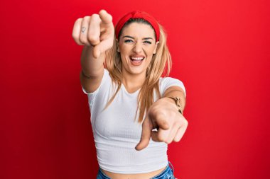 Beyaz tişörtlü genç beyaz kadın seni ve kamerayı işaret ediyor, pozitif ve neşeli gülümsüyor. 