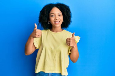 Genç Afro-Amerikan kızı, günlük kıyafetler giyiyor, başarı işareti yapıyor, elleriyle olumlu jest yapıyor, gülümsüyor ve mutlu. Neşeli ifade ve kazanan jest. 