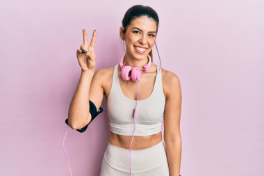 Spor kıyafetleri giyen ve kulaklık takan genç İspanyol kadın iki numarayı gösterip işaret ederken kendinden emin ve mutlu bir şekilde gülümsüyor.. 