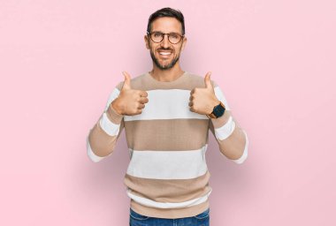 Sakallı, günlük kıyafetler giyen, gözlüklü, başarı işareti yapan, el hareketi yapan, gülümseyen ve mutlu bir adam. Neşeli ifade ve kazanan jest. 