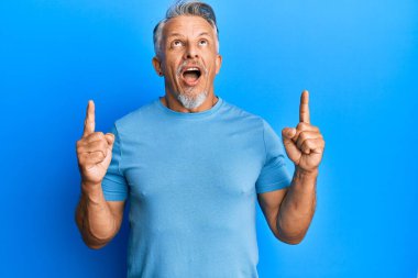 Orta yaşlı, gri saçlı, sıradan kıyafetler giyen, hayrete düşmüş, şaşırmış, parmaklarıyla işaret eden ve kollarını kaldıran bir adam.. 