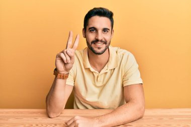 Latin asıllı genç adam, gündelik giysiler içinde masada oturuyor ve iki numaralı parmaklarıyla işaret ederek gülümsüyor ve mutlu.. 