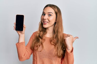 Akıllı telefon tutan genç sarışın kadın ekranı işaret ediyor başparmağını yukarı kaldırıyor ve ağzı açık gülümsüyor. 