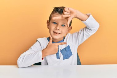 Sevimli beyaz çocuk, günlük giysiler giymiş masaya oturmuş gülümsüyor ve elleri ve parmakları mutlu yüzlü bir çerçeve yapıyor. yaratıcılık ve fotoğrafçılık kavramı. 