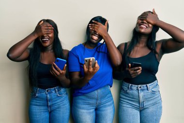 Üç Afro-Amerikalı genç arkadaş akıllı telefon kullanıyor. Gülümsüyor ve gülümsüyor. Gözleri şaşkınlık için kapatıyorlar. kör kavram. 