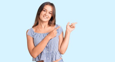 Güzel, beyaz bir kadın, günlük kıyafetler giymiş, gülümseyen ve kameraya bakıp iki elini ve parmağını yana doğru gösteren.. 