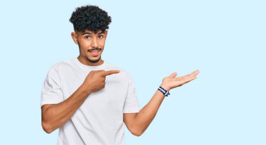 Gündelik beyaz tişört giyen genç Arap adam kameraya hayret ediyor ve gülümsüyor.. 