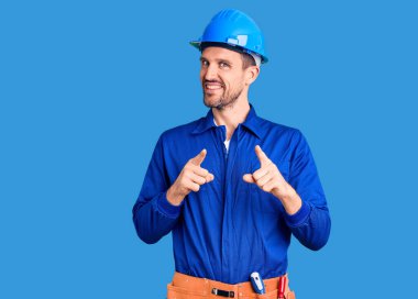 Genç, yakışıklı, işçi üniforması giyen ve şapkalı bir adam kamerayı mutlu ve komik yüzüyle işaret ediyor. iyi enerji ve titreşimler. 