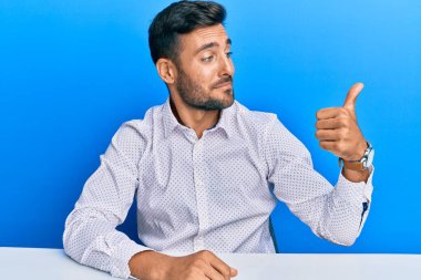İş kıyafetleri giymiş yakışıklı İspanyol bir adam masaya oturmuş gururlu görünüyor, gülümsüyor yan tarafa baş parmak işareti yapıyor. 