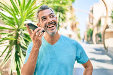 Orta yaşlı, kır saçlı, akıllı telefondan konuşan bir adam şehrin caddesinde yürüyor..