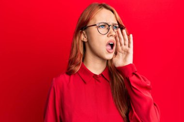 Genç, güzel, kızıl saçlı bir kadın kırmızı arka planda günlük kıyafetler ve gözlük takıp el ele bağırıyor ve çığlık çığlığa bağırıyordu. iletişim konsepti. 