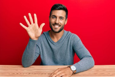 Yakışıklı İspanyol bir adam, sıradan giyinmiş, masanın üzerinde oturuyor ve beş numarayı işaret ediyor, kendinden emin ve mutlu gülümsüyor.. 