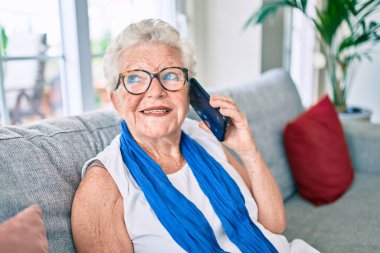 Yaşlı, yaşlı, gri saçlı, gülümseyen, mutlu bir kadın. Evdeki kanepede oturmuş telefonla konuşuyor.