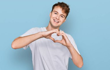 Gündelik beyaz tişört giyen genç beyaz bir adam aşıkken gülümsüyor elleriyle kalp sembolü yapıyor. romantik kavram. 