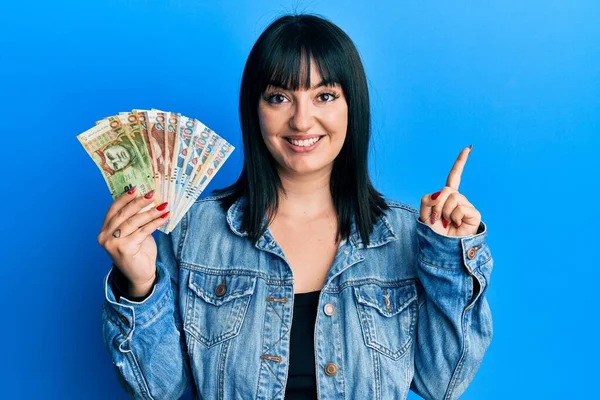 Young hispanic woman holding peruvian sol banknotes smiling happy pointing with hand and finger to the side