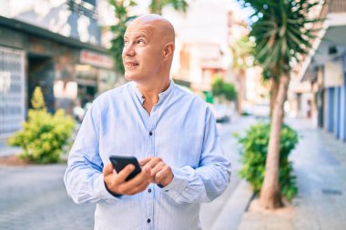 Orta yaşlı kel adam şehirde akıllı telefon kullanarak mutlu bir şekilde gülümsüyor..