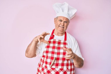 Kıdemli, gri saçlı, profesyonel fırıncı önlüğü giyen, tiksindirici, tiksindirici ve tiksindirici bir ifade takınan bir adam. Eller havada. 