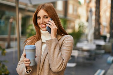 Genç kızıl saçlı kız akıllı telefonda konuşuyor. Şehirde kahve içiyor..