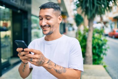 Şehrin caddesinde akıllı telefon kullanan, mutlu gülümseyen genç İspanyol adam..